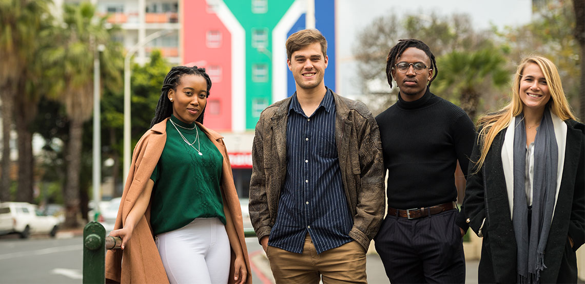A group of young South Africans pose smiling before a building emblazoned with the colors of South Africa's flag.