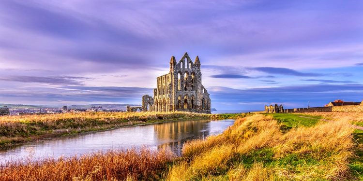 Whitby Abbey, a church in ruins across a river with purple sky