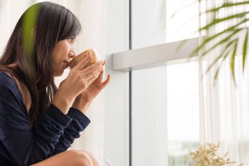 young woman sipping from a mug while looking out a bright window
