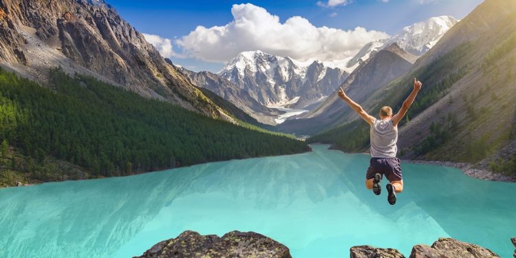 young man jumping in the air in front of blue water and snow-capped mountains