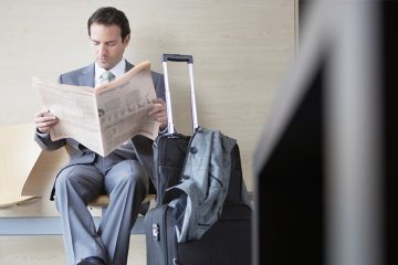 man in business attire reading a newspaper with luggage nearby