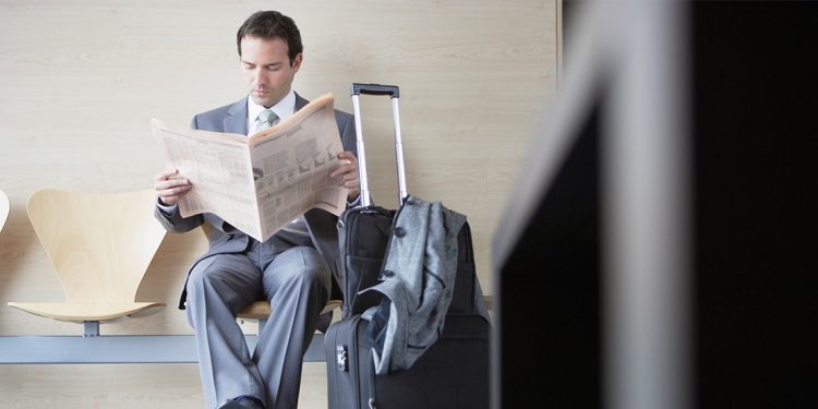 man in business attire reading a newspaper with luggage nearby