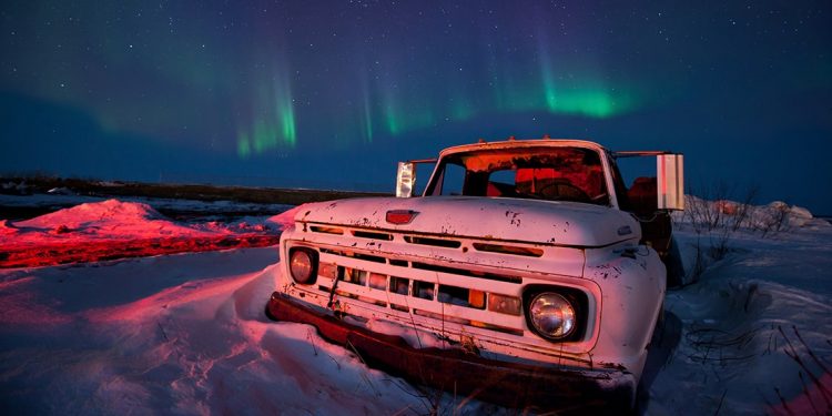An old trucked parked in the snow with green Aurora Borealis lights in the sky beyond