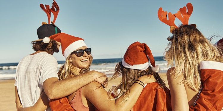Group of smiling young people on a beach wearing Santa hats and reindeer antlers