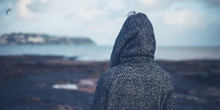 Person in a hooded sweatshirt stares looks out over a rocky beach