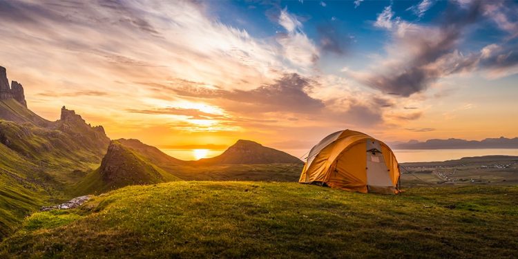 Sun rises over the water with a tent pitched on grass in the foreground