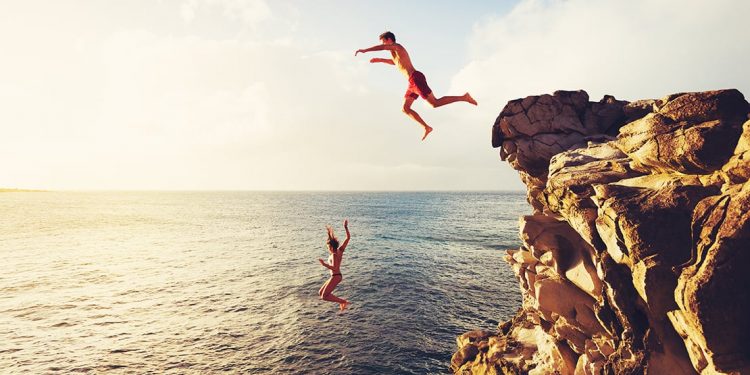 A man and woman jump off tall rocks into the ocean