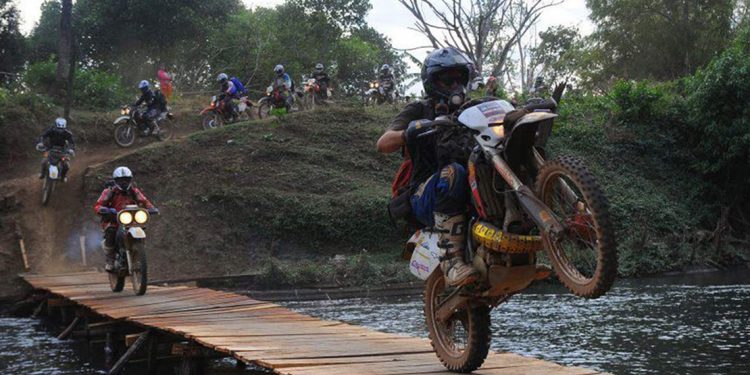 A row of motorcyclists ride down a hill and onto a rickety boardwalk over water