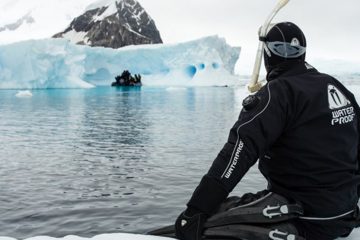 Person in wetsuit and scuba gear sits on an ice shelf with their feet in the water