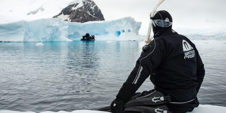 Person in wetsuit and scuba gear sits on an ice shelf with their feet in the water