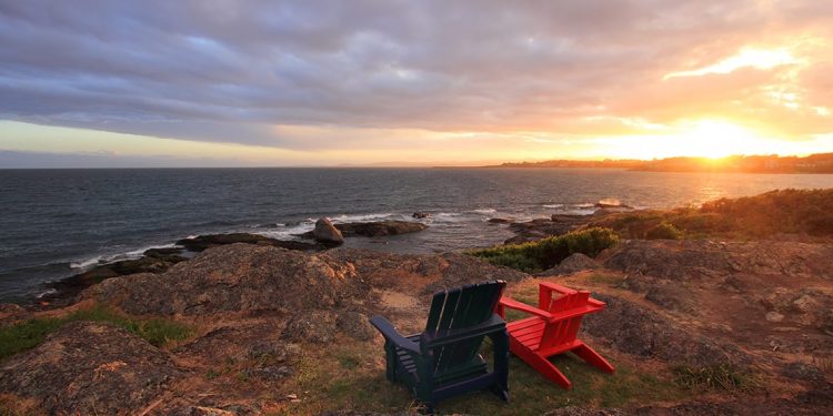 Two Adirondack chairs sit facing the ocean as the sun sets