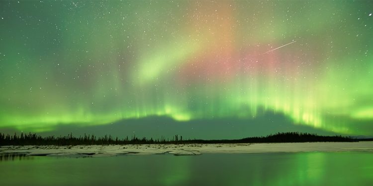 The wintery landscape of Alaska's country side shines of green from the Northern Lights and a shooting star makes an appear