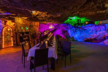 A large dining table is surrounded by colorful blue, purple and green lighting, in a cave setting