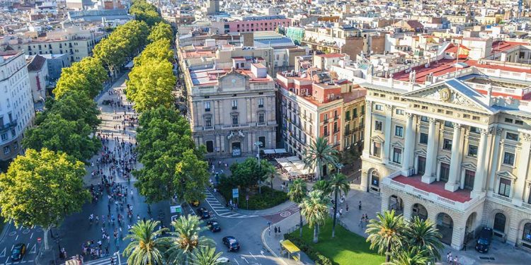 An aerial view of Barcelona, Spain includes old brick architectural buildings, many of the buildings include red roofing, and the streets are filled with people walking along large sidewalks