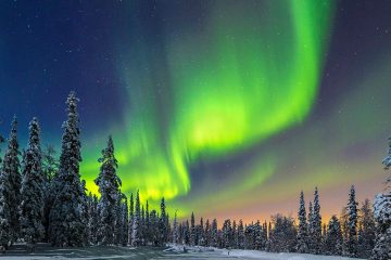 The Northern Lights show colors of green, orange, and red around snow-covered cedar trees