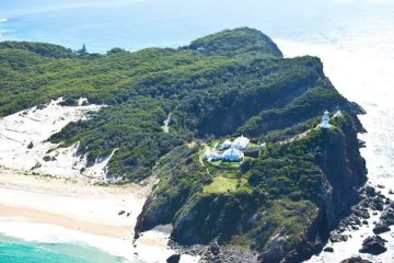 A tiny coastal island has four houses and a lighthouse, and is surrounded by ocean, nature, and beaches with white sand