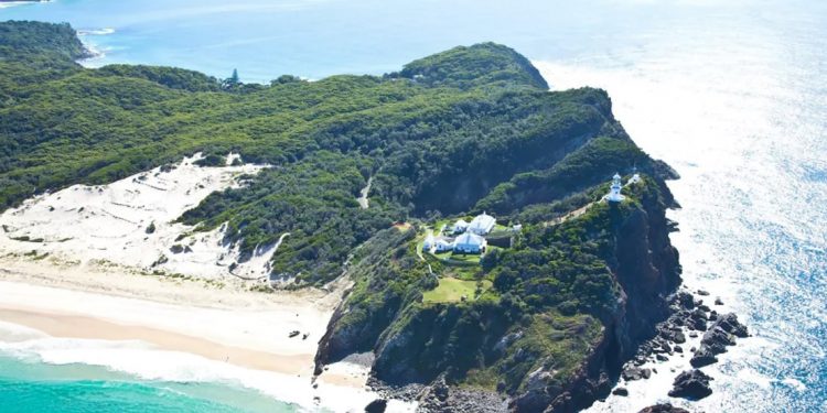 A tiny coastal island has four houses and a lighthouse, and is surrounded by ocean, nature, and beaches with white sand
