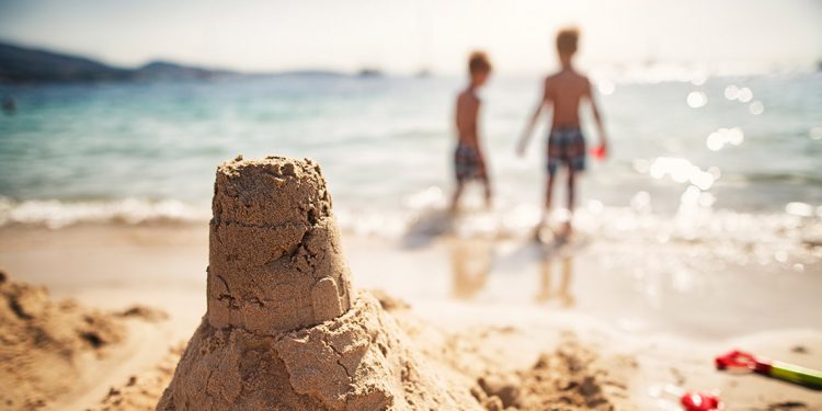 A handmade sand castle is in focus with two young children in the background