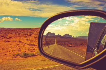 A car's side mirror looks back on the road while travelling with the desert in the background