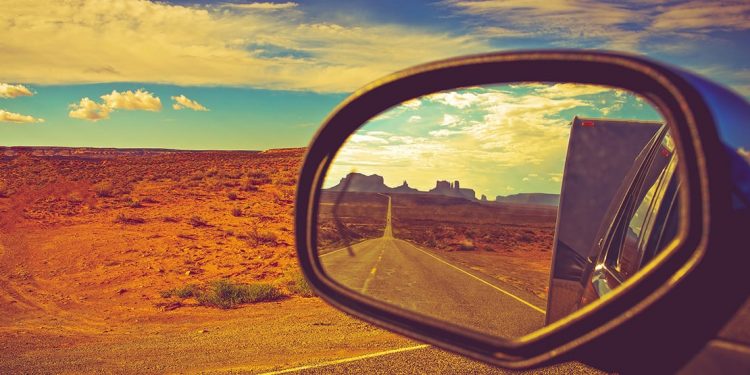 A car's side mirror looks back on the road while travelling with the desert in the background