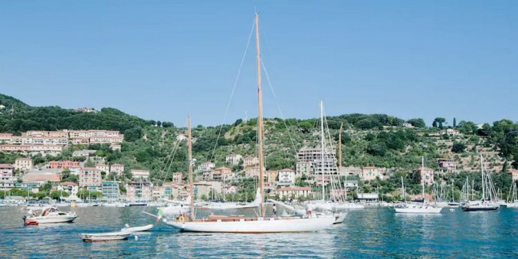 A restored 60-foot yacht built in 1948 with colors of white and natural wood sails along the coastline of Italy
