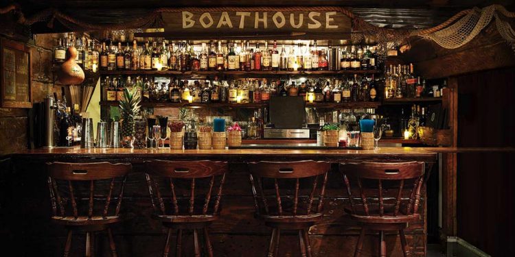 Four bar stools sit alongside a bar table and in the background is shelves of alcohol and a sign that says "boathouse"