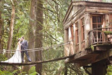 A bride and a groom are kissing on a bridge that attaches to a tree house in the woods