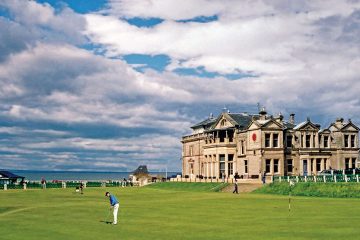 The famous St. Andrew's building in Scotland is in the background while a golfer is teeing off on the golf course