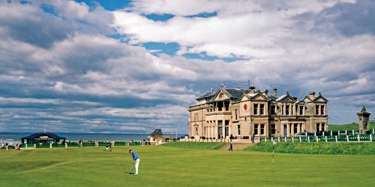 The famous St. Andrew's building in Scotland is in the background while a golfer is teeing off on the golf course