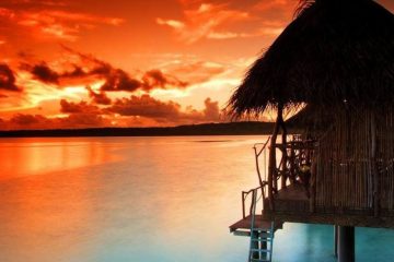 Hut with thatched roof on stilts over the water with fiery red sunset on the horizon.