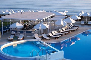 White umbrellas along the beach by the ocean. Concrete deck around a pool right next to beach with raised hot tub.