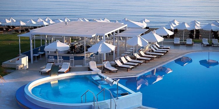 White umbrellas along the beach by the ocean. Concrete deck around a pool right next to beach with raised hot tub.