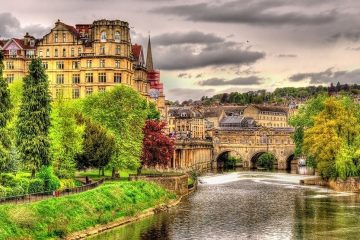 River leading to stone bridge with greenery on either side. Town buildings visible on other side of bridge.