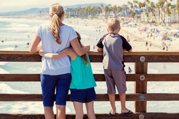 Daughter has arms wrapped around her mom's waist as they look out at a beach from up on a boardwalk. Son is standing on bottom rung of fence beside them.