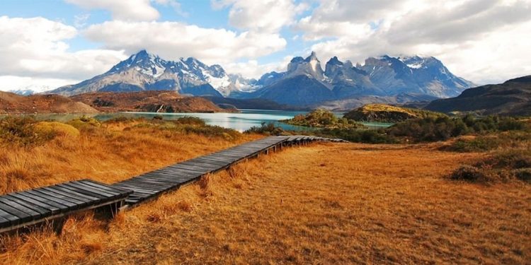 Boardwalk leading out across yellowed grasses, down to lake with mountains in distance.
