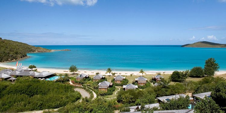 Blue ocean with strip of white sand beach and various private huts on green grass surrounded by shrubbery.