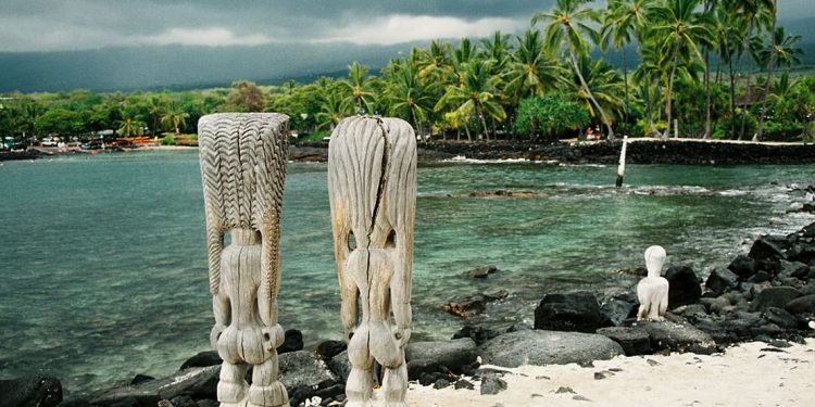 Two wooden tiki statues at the edge of the ocean with black rocks and palm trees. Skies look stormy.