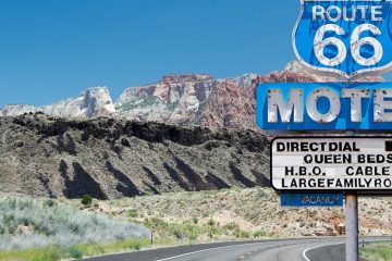 Blue roadside sign for Route 66 motel in front of curve of road with mountain rising up on left side.