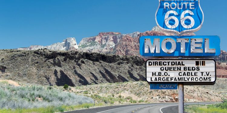 Blue roadside sign for Route 66 motel in front of curve of road with mountain rising up on left side.