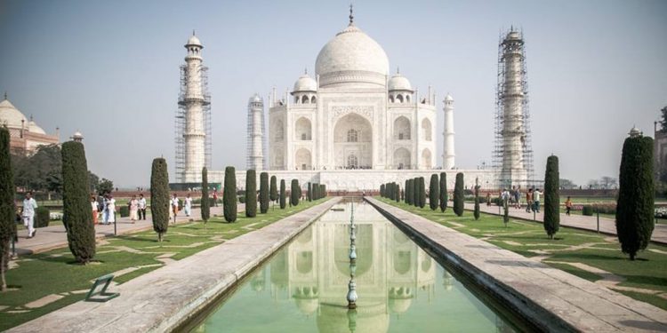 A view from the rectangular pool in front of building. Reflection of Taj Mahal is visible in the water. There are tall, carefully groomed trees on either side of pool.