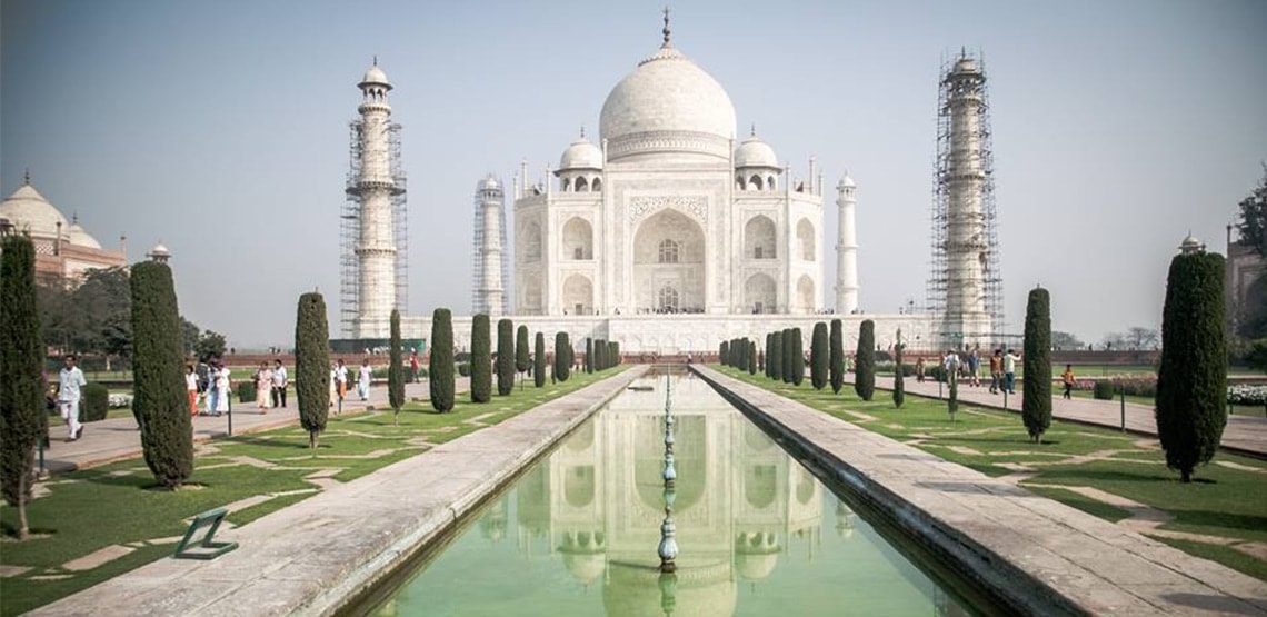 A view from the rectangular pool in front of building. Reflection of Taj Mahal is visible in the water. There are tall, carefully groomed trees on either side of pool.