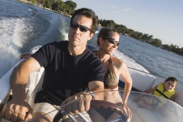 Middle aged man driving speed boat with wife in bikini seated to his left and child wearing yellow life jacket on set behind them.
