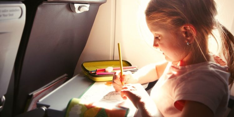 Little girl on airplane with tray down, drawing on piece of paper with pencil case open beside her.
