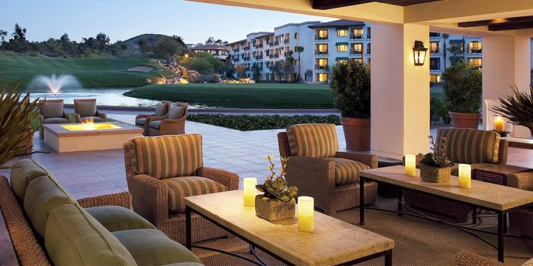 Outdoor patio with lounge chairs, rectangular tables with candles and centerpieces, and a fountain. Golf course in the background.