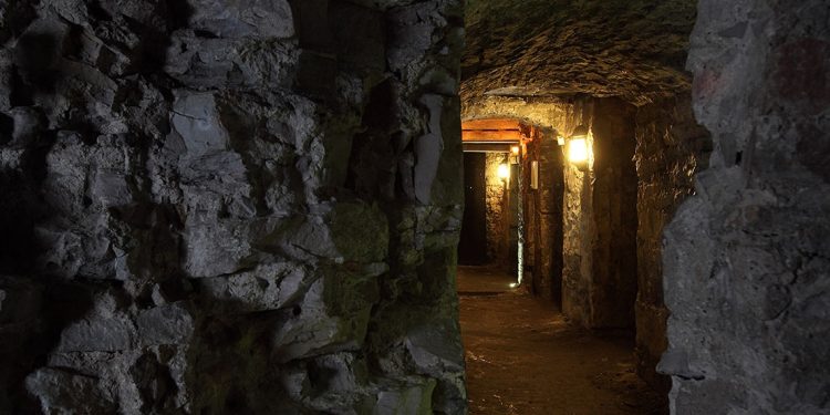 Dark tunnel with uneven stone walls and lanterns lining the walls.