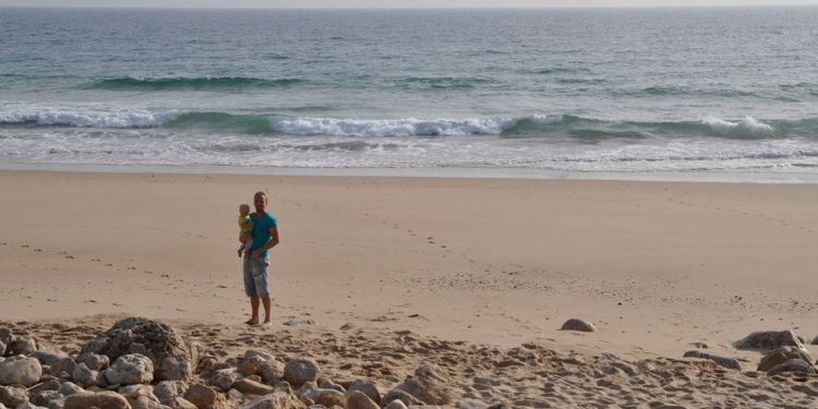 Man holding toddler on a beach in front of waves. Rocks in front of him.