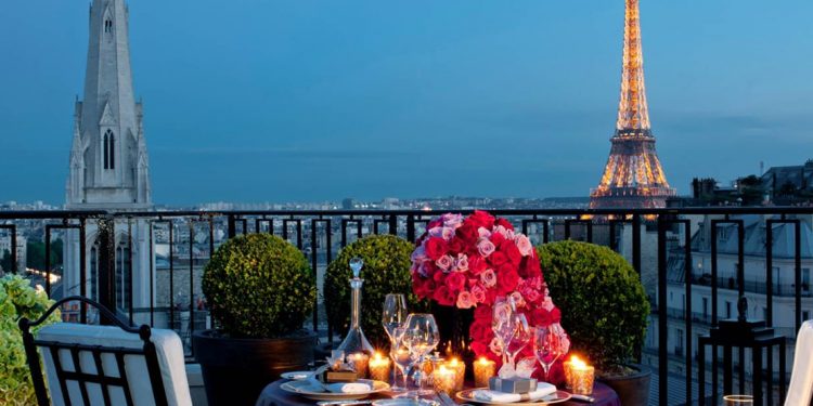 Balcony with candlelit dinner on small table with Eiffel Tower and church spire visible over railing.
