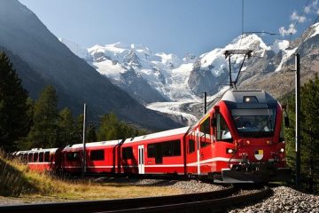 Red train coming around a corner with white peaked mountains in the background.