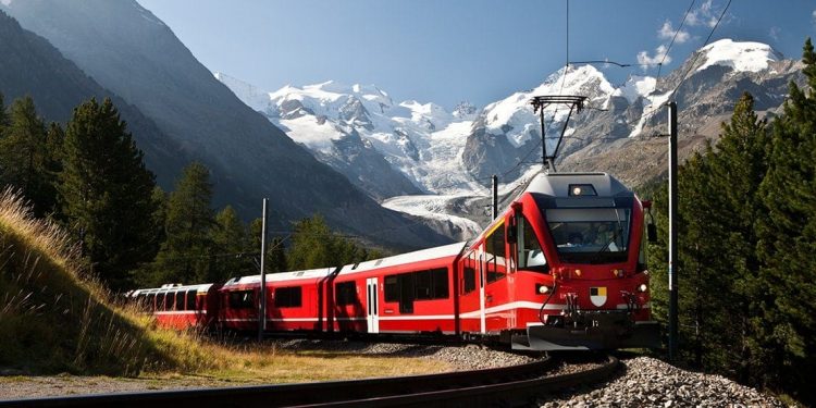 Red train coming around a corner with white peaked mountains in the background.