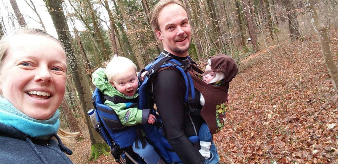 Mom on left, dad with baby in carrier on front and toddler in carrier on back to her right. In woods with leaves on ground.
